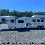 White horse trailer with two axles and side windows on a gravel lot, backed by a tree line and clear blue sky.