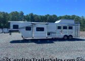 White horse trailer with two axles and side windows on a gravel lot, backed by a tree line and clear blue sky.
