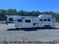 White horse trailer with two axles and side windows on a gravel lot, backed by a tree line and clear blue sky.