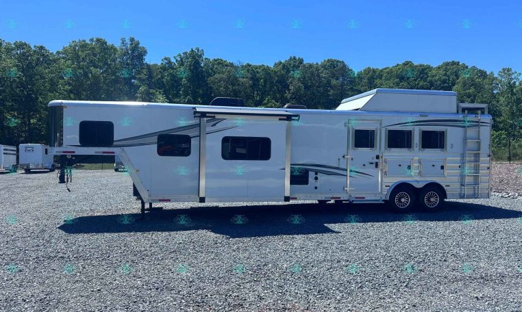 White horse trailer with two axles and side windows on a gravel lot, backed by a tree line and clear blue sky.