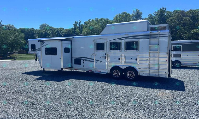 White fifth-wheel RV trailer parked on a gravel lot with trees in the background; dealership watermark at bottom.