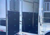 Open side of a white horse trailer with doors swung wide, showing interior partition and wall hooks for tack on a black lower wall edge.