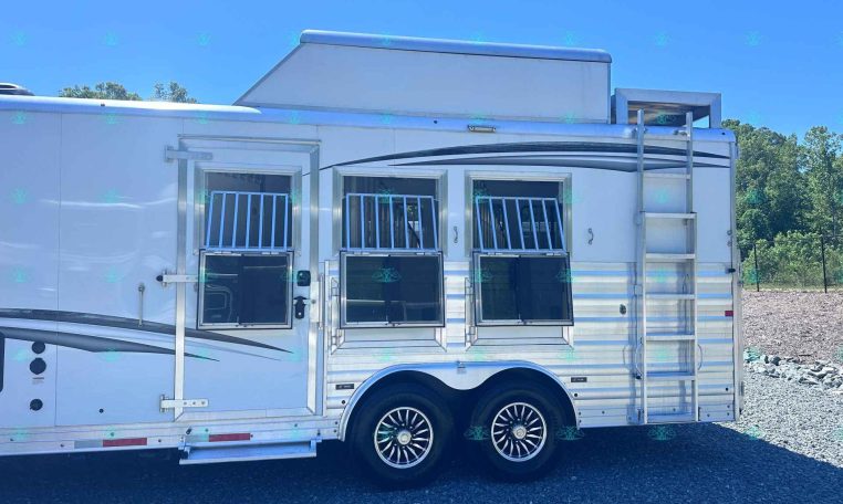 White horse trailer with barred windows and chrome wheels, parked on gravel under a clear blue sky.
