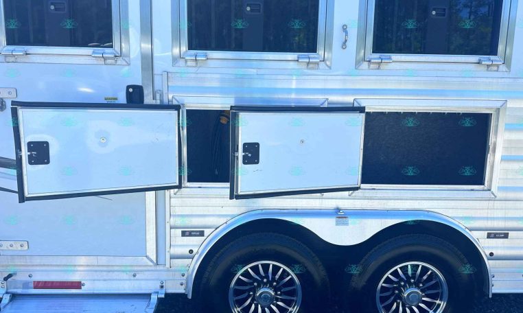 Side view of a silver trailer with open storage compartments and chrome wheels; CarolinaTrailerSales.com visible at the bottom.