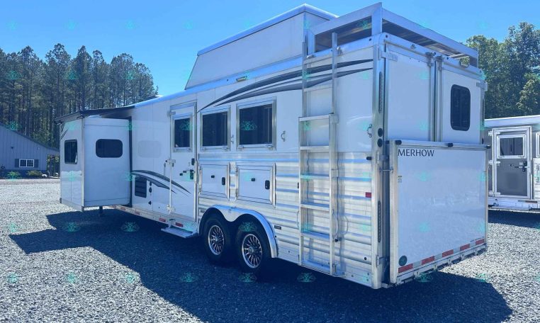 White horse trailer with an open side door and ladder, parked on a gravel lot with trees in the background.