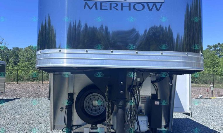 Front view of a blue Merhow horse trailer with its logo, parked on a gravel lot under a clear blue sky