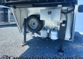 Open trailer rear storage compartment containing a mounted spare tire, propane tanks, and a metal toolbox/box with wiring nearby for utilities of the trailer