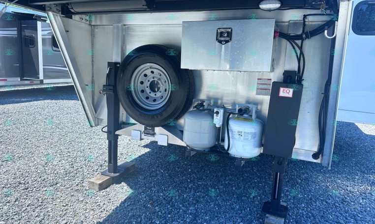 Open trailer rear storage compartment containing a mounted spare tire, propane tanks, and a metal toolbox/box with wiring nearby for utilities of the trailer