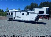 White fifth-wheel RV trailer with a raised front cab, side entry door, and long gray decorative graphics, parked on a gravel lot with trees and blue sky in the background.