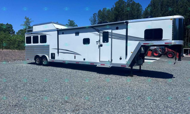 White fifth-wheel RV trailer with a raised front cab, side entry door, and long gray decorative graphics, parked on a gravel lot with trees and blue sky in the background.