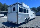 White Merhow horse trailer with windows and aluminum siding parked on a gravel lot, trees in the background.