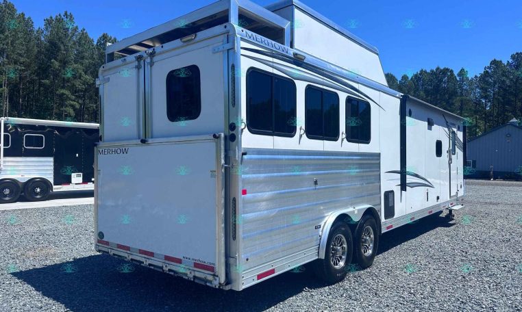 White Merhow horse trailer with windows and aluminum siding parked on a gravel lot, trees in the background.