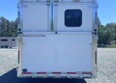 Rear view of a white Merhow horse trailer with double doors and a small rounded window, parked on gravel under a blue sky.