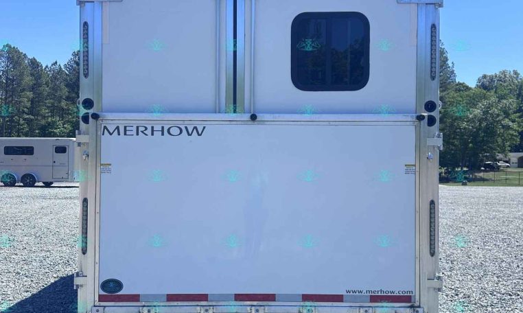 Rear view of a white Merhow horse trailer with double doors and a small rounded window, parked on gravel under a blue sky.