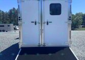 Rear view of a white trailer with a lowered black loading ramp on a gravel lot, trees in the background under a clear blue sky.