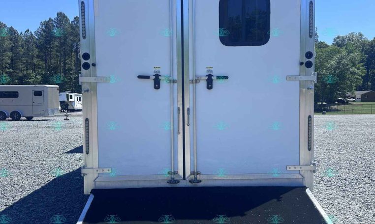 Rear view of a white trailer with a lowered black loading ramp on a gravel lot, trees in the background under a clear blue sky.