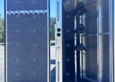 Open aluminum trailer with the door swung open, showing a dark interior and a vertical rack/seat structure along the left side; exterior is on a gravel lot under a clear blue sky.