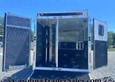 Open horse trailer with ramp down, showing interior stalls and divider on a gravel lot under a blue sky.
