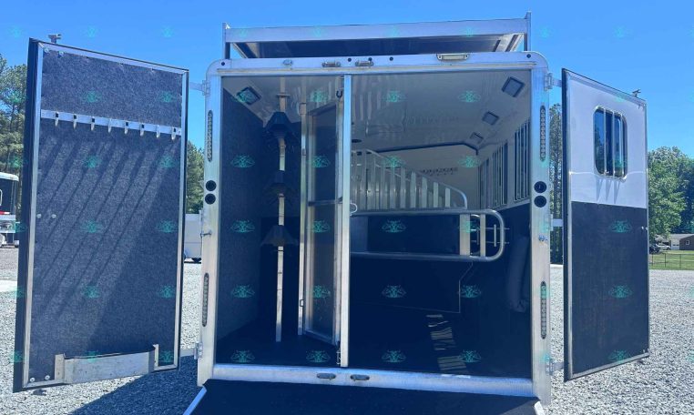 Open horse trailer with ramp down, showing interior stalls and divider on a gravel lot under a blue sky.