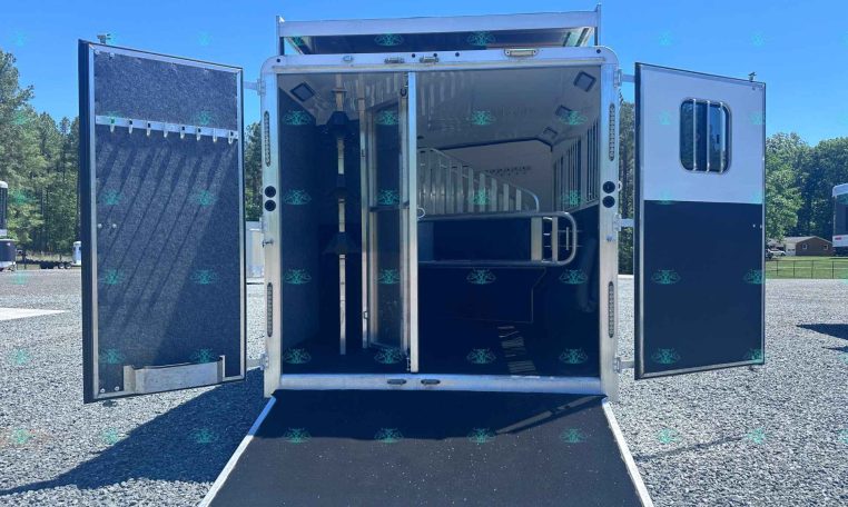 Open aluminum horse trailer with ramp down, interior stalls and tie rails visible in a sunny outdoor lot.