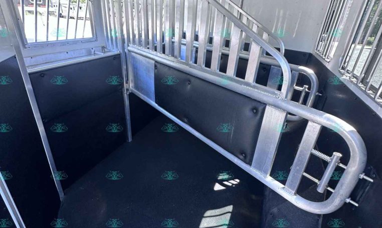 Interior of a silver horse trailer showing a central divider, aluminum rails, and window bars along the walls for ventilation and separation of stalls.