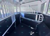Interior of a horse trailer with two metal stalls and padded lower walls, partitions between stalls, and hooks along the wall for tack storage.