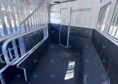 Interior of a white horse trailer showing two stalls with metal dividers, padded black walls, and a central doorway between stalls.