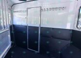 Interior of a light trailer with two rows of metal hooks along the walls and a central door, black cushioned lower walls, and windows visible