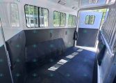 Interior of a livestock trailer with metal-barred windows and padded side walls; a doorway at the far end leads outside to green trees.