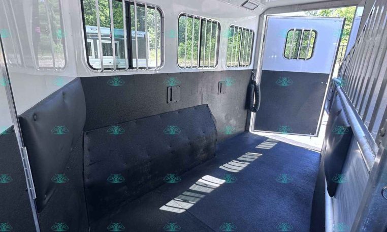 Interior of a livestock trailer with metal-barred windows and padded side walls; a doorway at the far end leads outside to green trees.