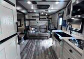 Bright RV interior with white cabinetry, gray sofa, and wood-look flooring across the space.