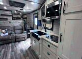 Modern RV kitchen and living area: white cabinets, stainless microwave, stove, sink, gray sofa, and wood-look flooring.