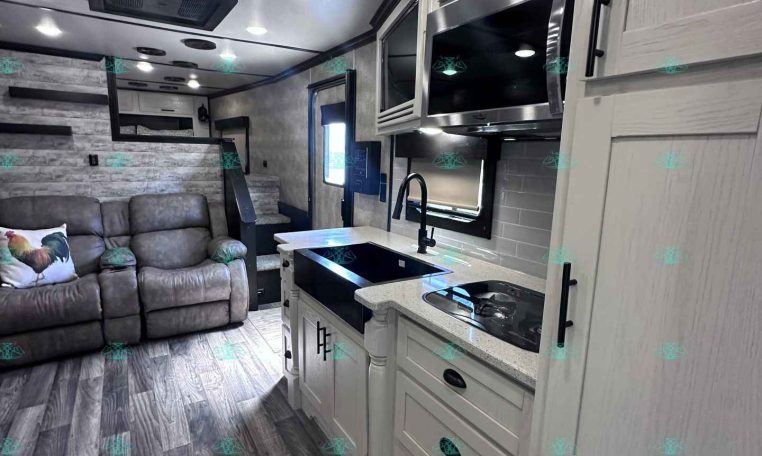 Modern RV kitchen and living area: white cabinets, stainless microwave, stove, sink, gray sofa, and wood-look flooring.