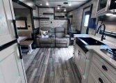 Interior of a modern travel trailer showing a kitchen with a white island and sink on the right, and a gray sofa with a decorative pillow in the living area.