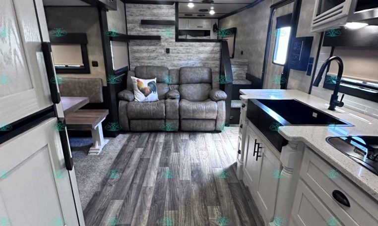 Interior of a modern travel trailer showing a kitchen with a white island and sink on the right, and a gray sofa with a decorative pillow in the living area.
