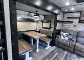Interior of a modern trailer living area with a wooden dining table, bench seating, and a gray sofa with a rooster pillow.
