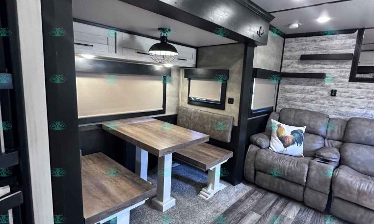 Interior of a modern trailer living area with a wooden dining table, bench seating, and a gray sofa with a rooster pillow.