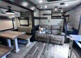 Cozy trailer interior with gray leather sofa, wooden dining table, and stairs to a loft area in a modern RV.