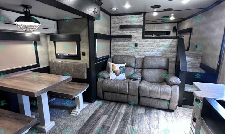 Cozy trailer interior with gray leather sofa, wooden dining table, and stairs to a loft area in a modern RV.