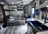 Open-plan RV interior with a gray sofa and rooster pillow, ceiling lights, and a compact kitchen island with a sink on the right.