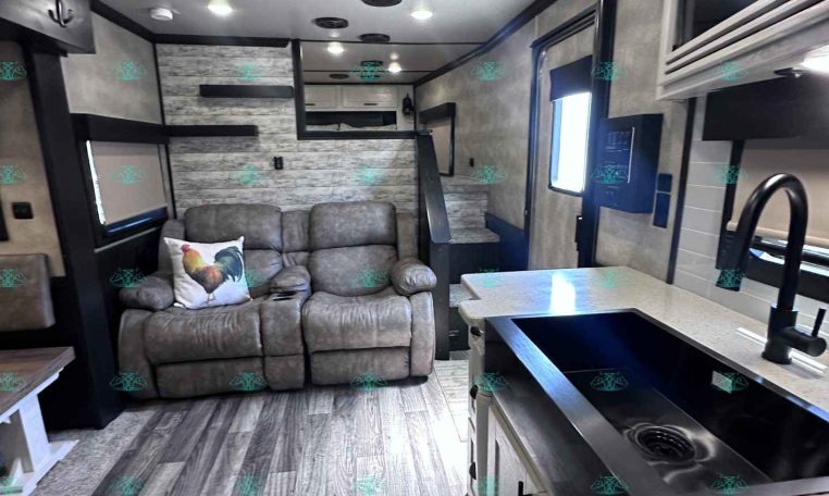 Open-plan RV interior with a gray sofa and rooster pillow, ceiling lights, and a compact kitchen island with a sink on the right.