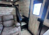 Interior of a compact travel trailer living area with a gray recliner sofa, light wood paneling, and a small staircase leading to a bunk area; a window with a blue frame and shade is on the right, and ceiling lights above.