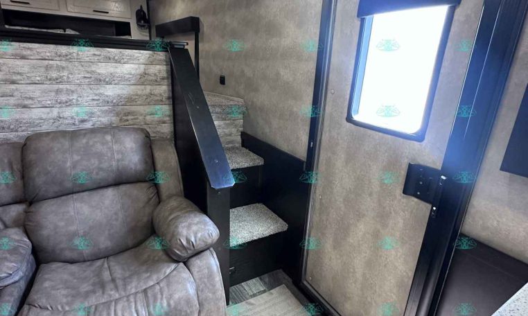Interior of a compact travel trailer living area with a gray recliner sofa, light wood paneling, and a small staircase leading to a bunk area; a window with a blue frame and shade is on the right, and ceiling lights above.