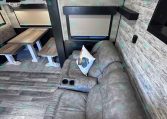 Cozy trailer living area with a gray sectional sofa, a wood dining table with benches, and wood-style walls.