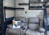 Cozy gray recliner sofa with a rooster pillow in a rustic trailer living area, wood panel walls and built-in shelves nearby.