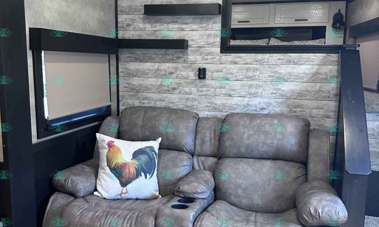 Cozy gray recliner sofa with a rooster pillow in a rustic trailer living area, wood panel walls and built-in shelves nearby.