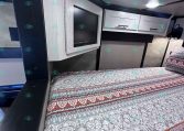 Interior of a travel trailer bedroom with a mounted flat-screen TV, white overhead cabinets, and a patterned bedspread in blue-gray tones.