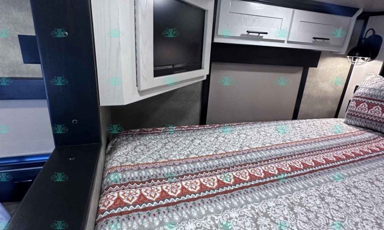 Interior of a travel trailer bedroom with a mounted flat-screen TV, white overhead cabinets, and a patterned bedspread in blue-gray tones.