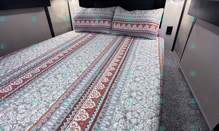 Bedroom inside a trailer with a patterned bedspread, two matching pillows, white overhead cabinets, and industrial-style wall lamps above the headboard edge of the bed is against the wall.