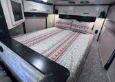 Interior of a travel trailer bedroom with a queen bed, patterned quilt, and overhead white cabinets above the headboard